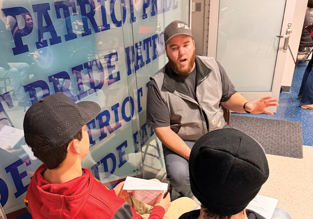 A bearded man wearing a gray vest and baseball cap talks animatedly to two young men sitting opposite him. One youth holds papers. A glass wall with repeating "PATRIOT PRIDE" text is in the background.