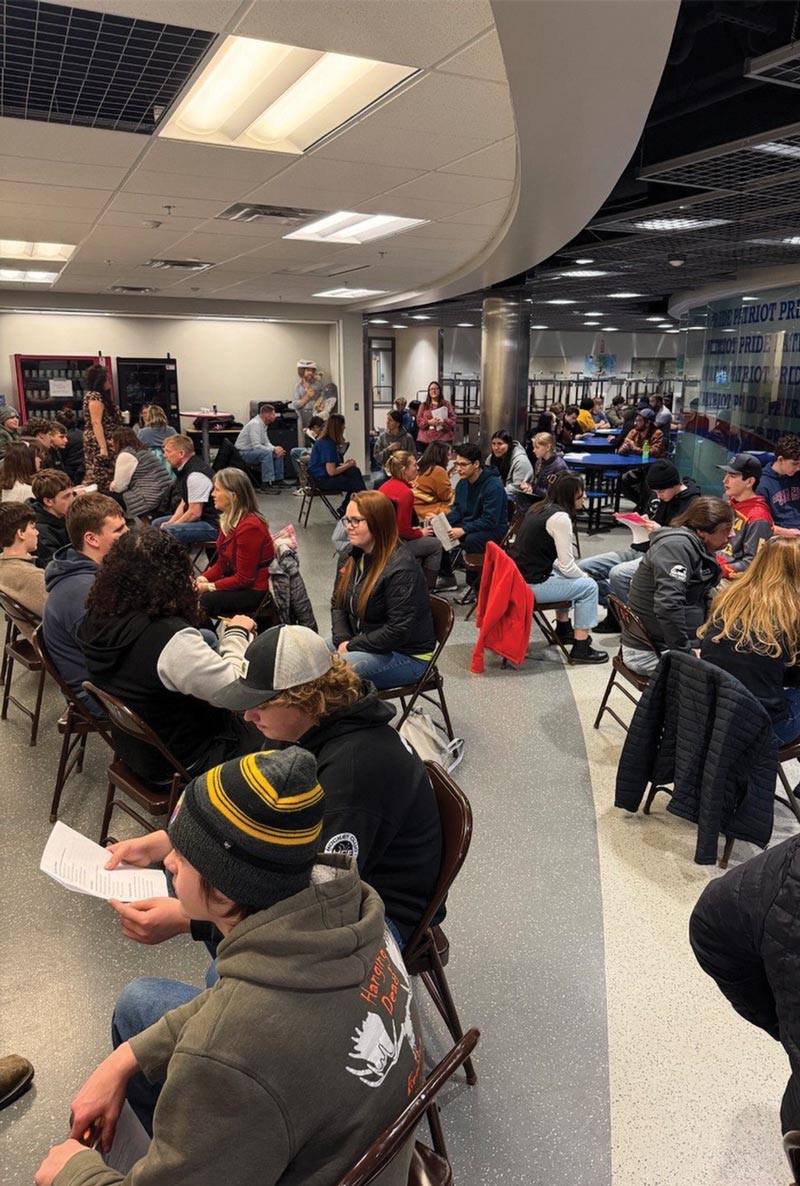 A long view of a hallway-like room with many people, adults and teens, sitting in small face-to-face clusters on folding chairs.