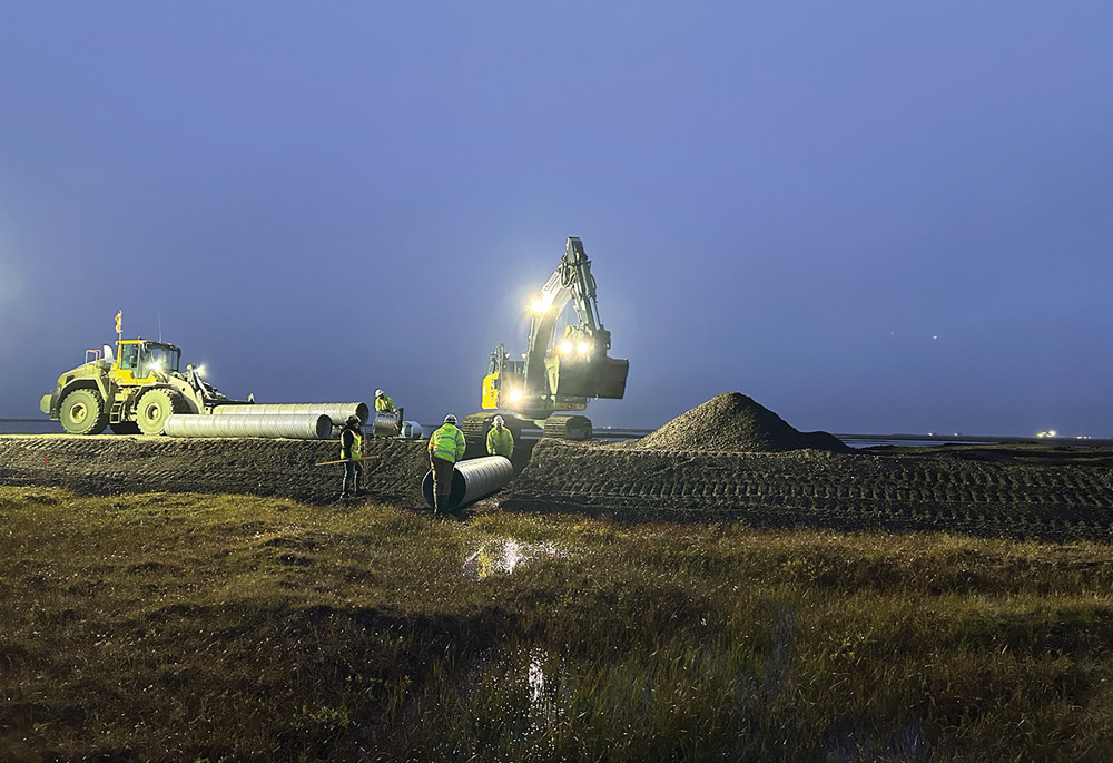 At night, construction workers in high-visibility vests use an excavator and front-end loader with bright lights to install a large metal culvert pipe into a trench. The site is surrounded by dark, marshy terrain.