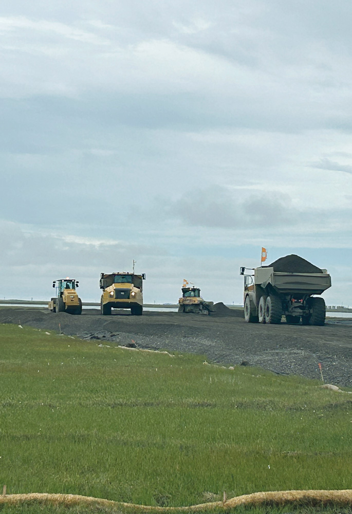 Heavy construction equipment, including a roller and haul trucks, work on a gravel surface next to a green field under a cloudy sky.