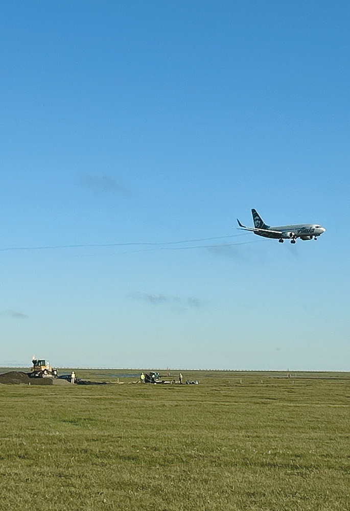 An Alaska Airlines passenger jet flies low over a grassy field where a bulldozer and several construction workers are visible in the distance.