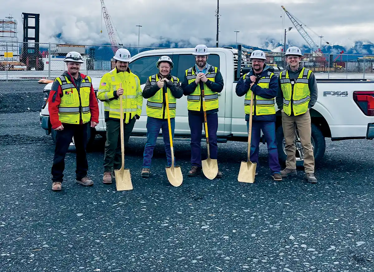 Six people in white hard hats and high-visibility safety vests shovels in front of a white pickup truck at an industrial construction site with misty mountains in the distance