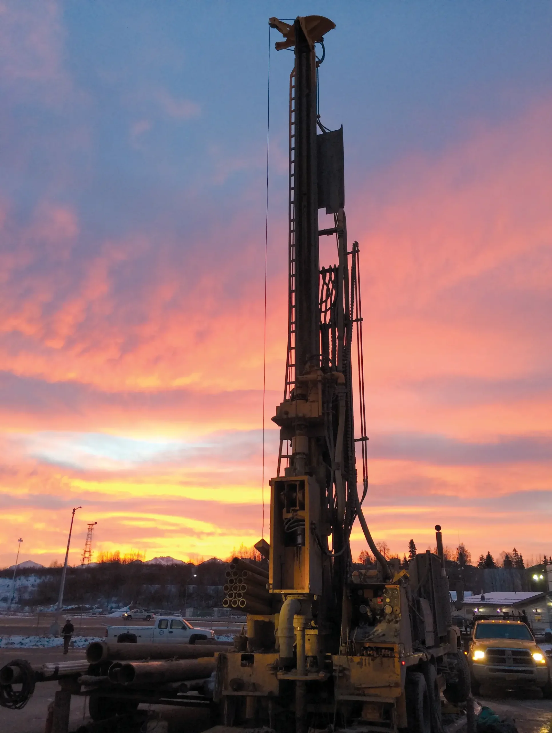 A tall industrial drilling rig silhouetted against a vibrant pink and orange sunset sky at a snowy Alaskan construction site