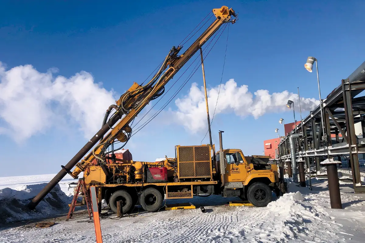 A yellow M-W Drilling truck-mounted rig with its mast extended diagonally into the ground at a snowy Alaskan industrial site