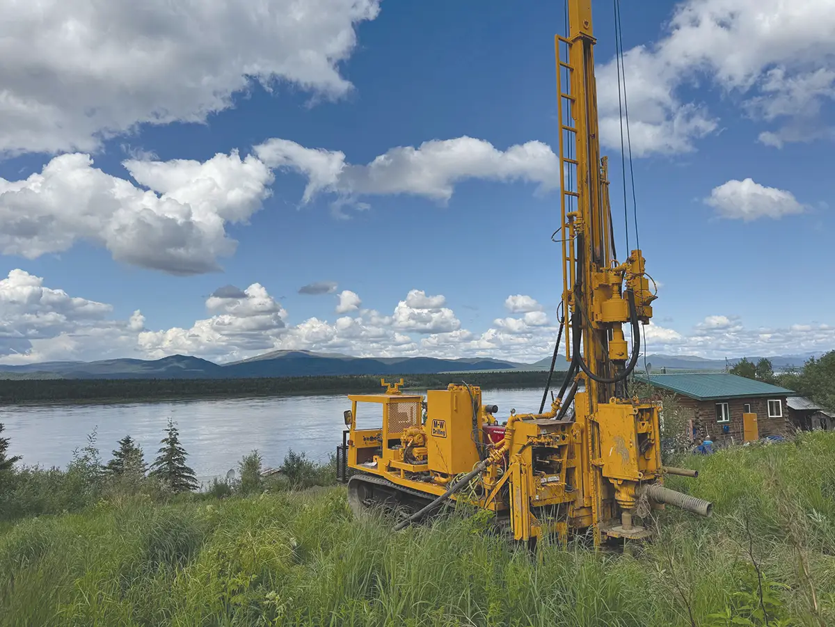 A large, yellow, track-mounted mobile drilling rig with its mast extended vertically, standing on a grassy bank overlooking a wide river with forested mountains under a partly cloudy sky