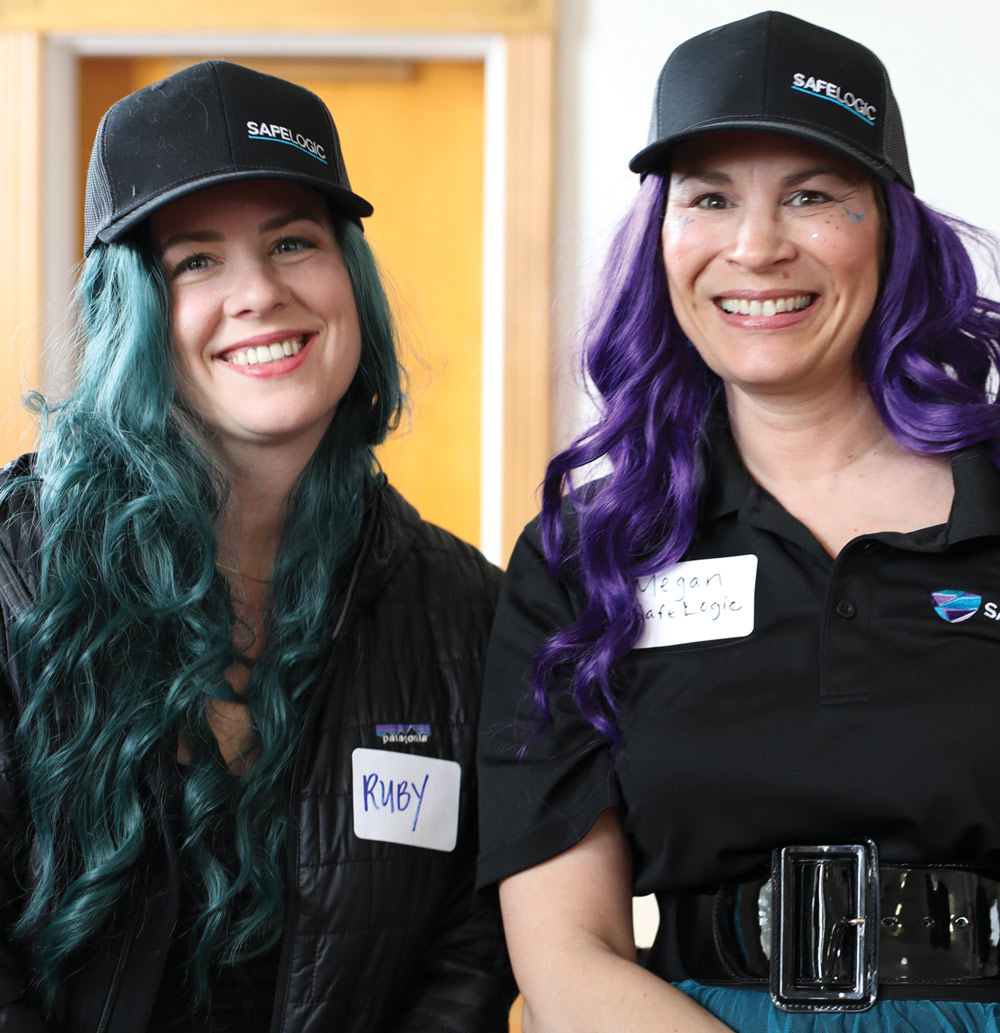 Ruby and Megan smile together wearing matching SafeLogic caps, showing off vibrant teal and purple hair.