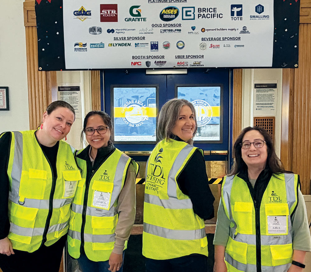 Four women wearing high-visibility TDL Staffing safety vests smile beneath a corporate sponsor banner.