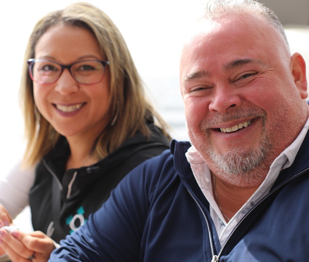 Close-up portrait of a smiling woman with glasses and a man in a navy zip-up smiling widely at an event.