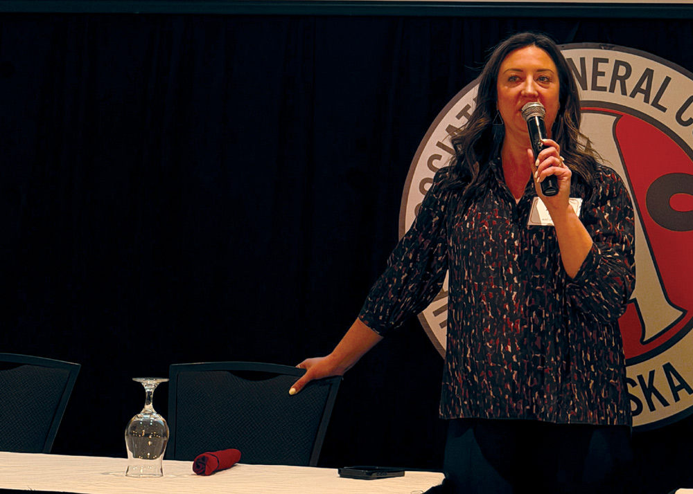 A woman stands at a table speaking into a microphone in front of a large circular AGC Alaska logo backdrop.