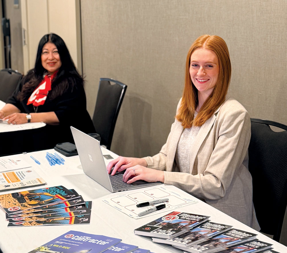 A smiling red-haired woman works on a laptop at a registration table filled with 2026 AGC member directories.