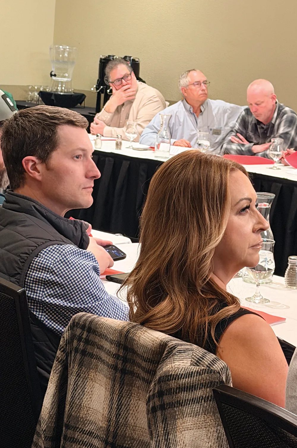 Event attendees sit at a table listening attentively, with a close-up profile view of a man and a woman in focus.