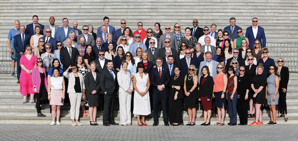 A large group of professionally dressed attendees poses together for a photo on wide stone steps outdoors.