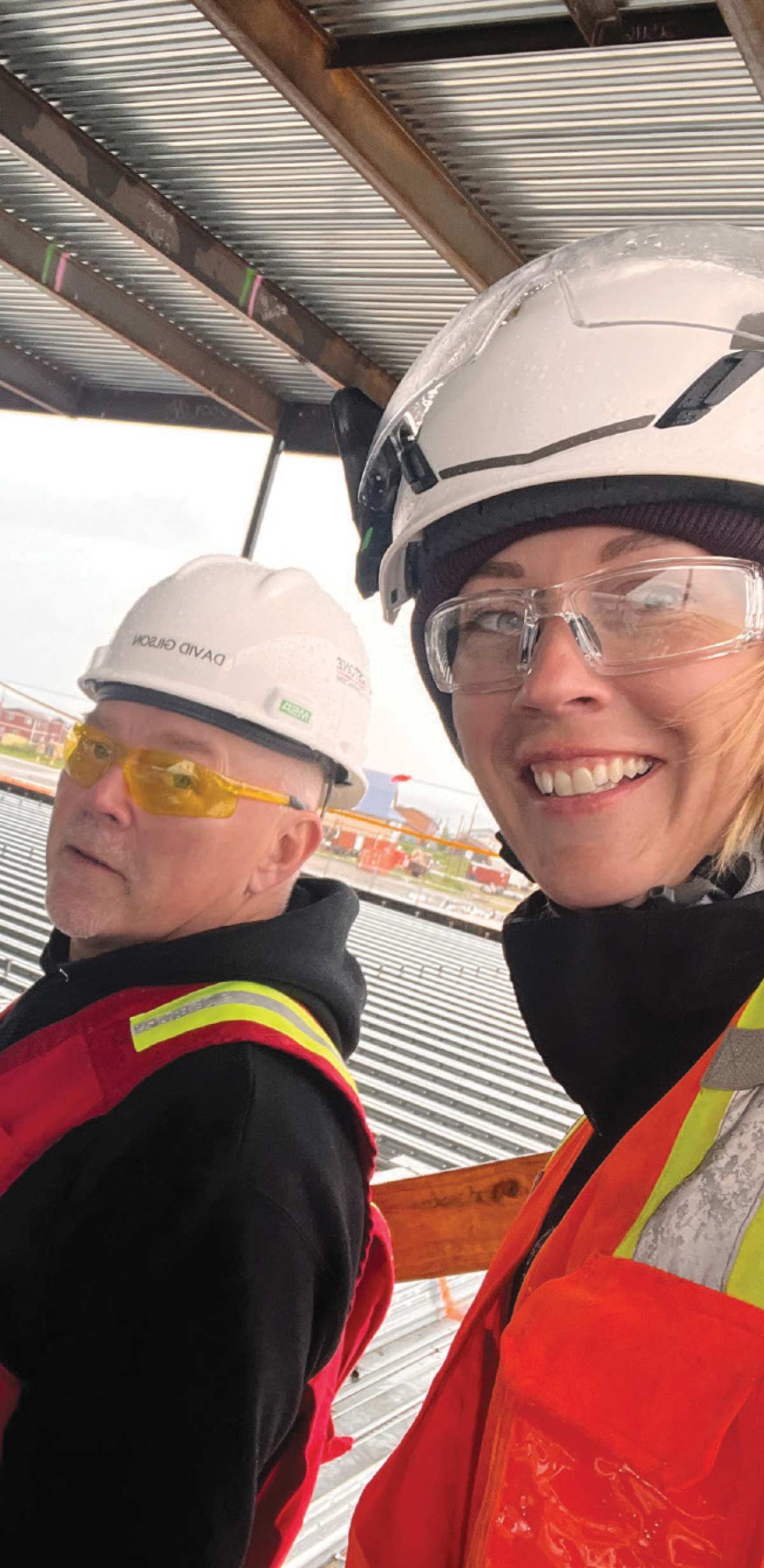 A close-up selfie of a smiling female construction worker in safety gear standing next to a male coworker beneath a corrugated metal ceiling grid.