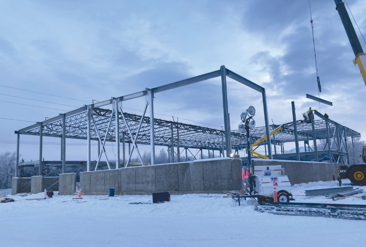 A large steel-framed building structure under construction stands in a snow-covered field under a cloudy, overcast winter sky.