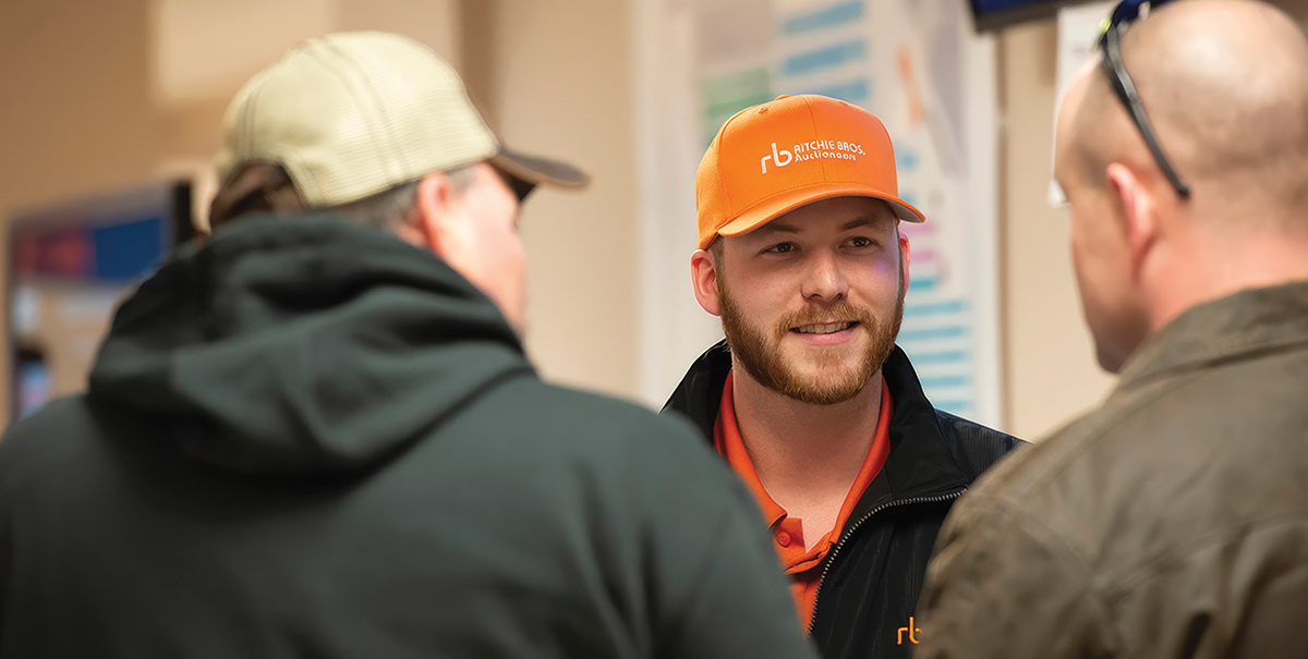 A bearded Ritchie Bros. staff member wearing an orange company cap and polo shirt smiles while speaking to two men in a close-up, over-the-shoulder view.