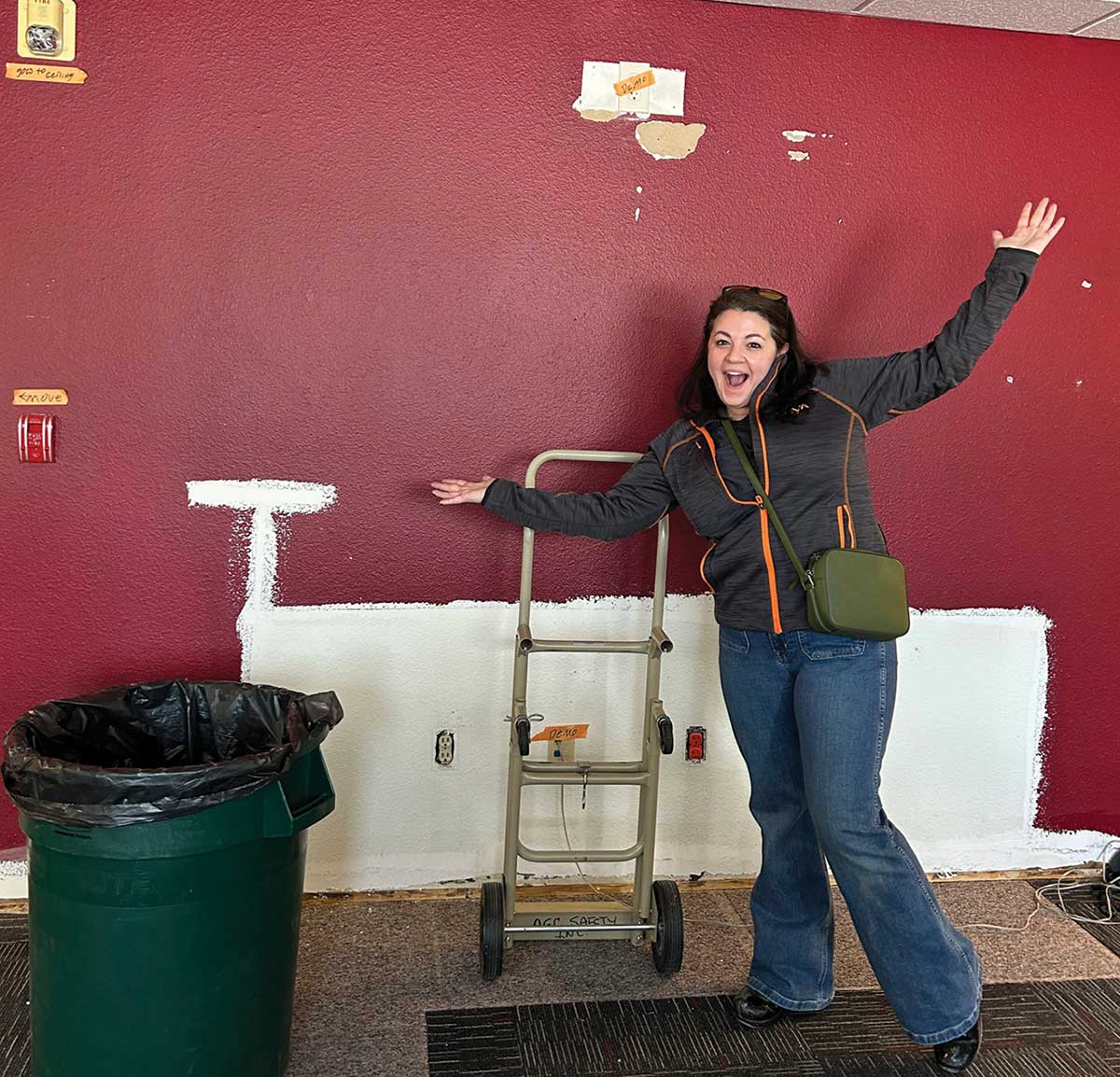 Catherine Sullivan stands with arms outstretched and a wide smile in front of a burgundy wall with a white painted outline. A green plastic trash can sits on the left and a metal hand truck is behind her.