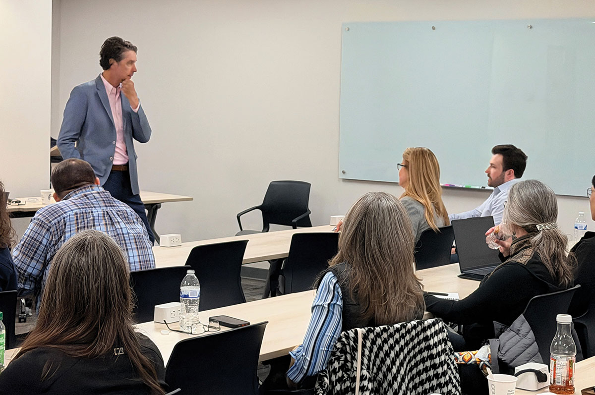 A man in a blue blazer stands at the front of a classroom with his hand on his chin, speaking to a group of seated attendees at long tables.