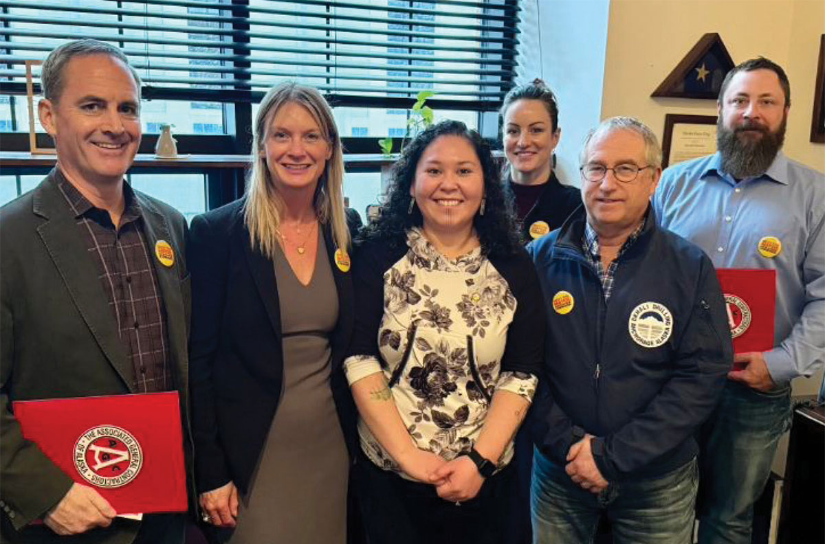 Six smiling people pose for a group photo in front of a window with blinds. Several individuals wear yellow event pins, and two men hold red folders with a circular AGC logo.