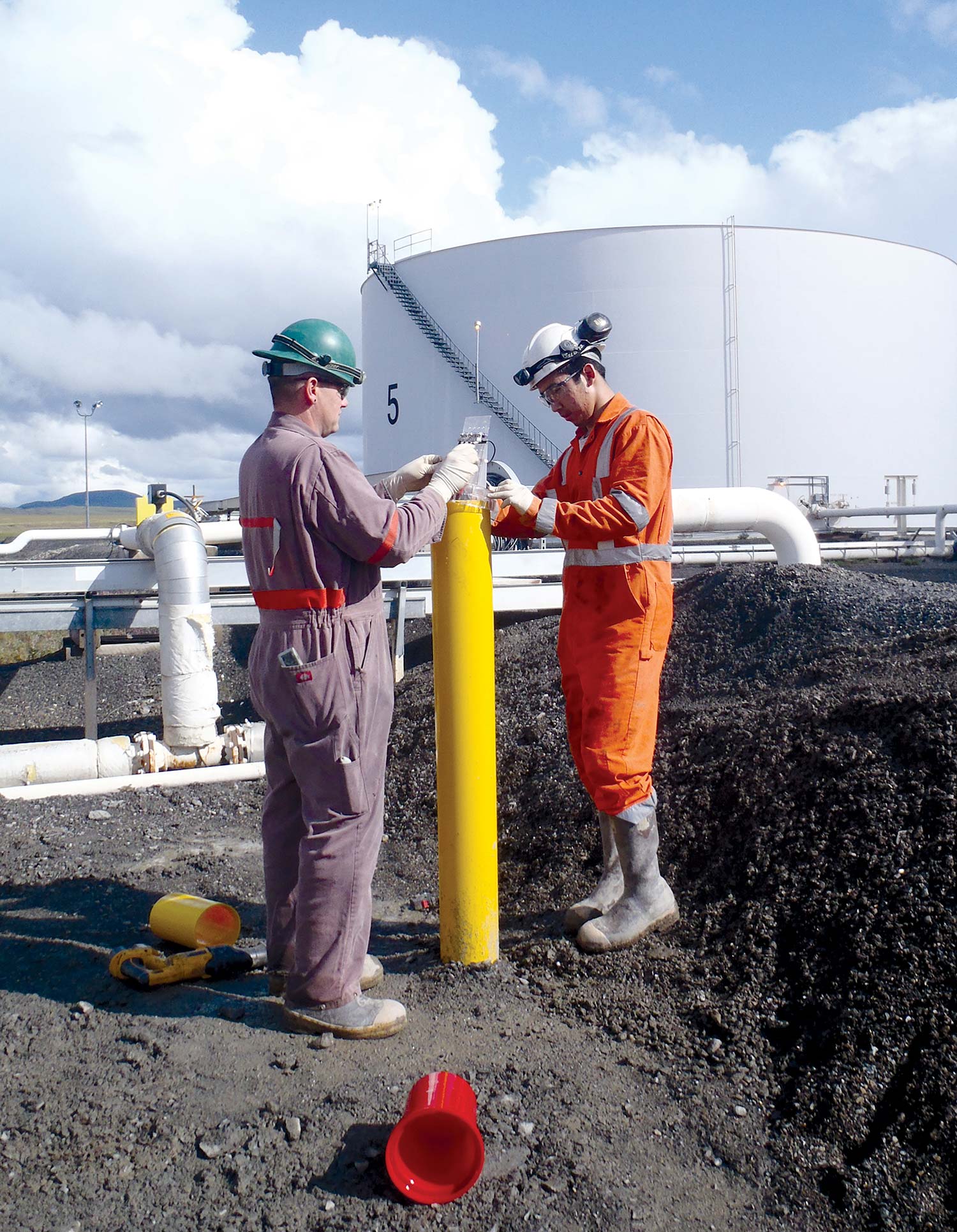 Two technicians working on a yellow pipeline structure near a large industrial storage tank.