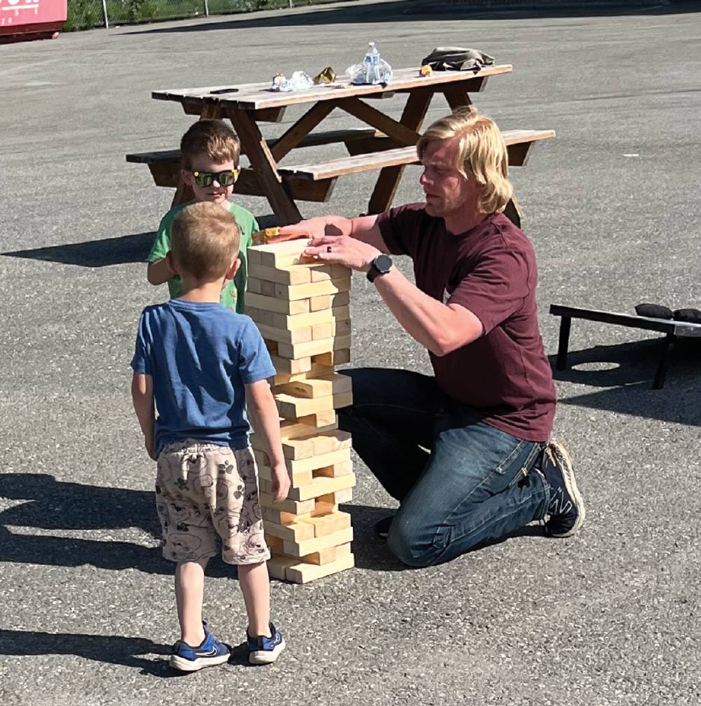 A man kneeling to play giant wooden block stacking game with two young boys outdoors.