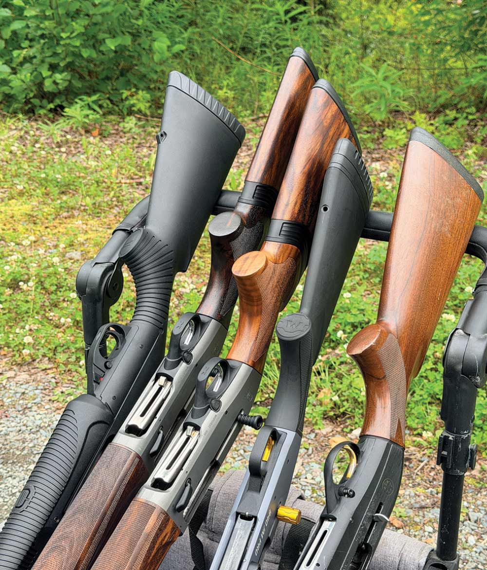 Close-up view of five shotguns with wood and synthetic stocks resting in a gun rack outdoors.