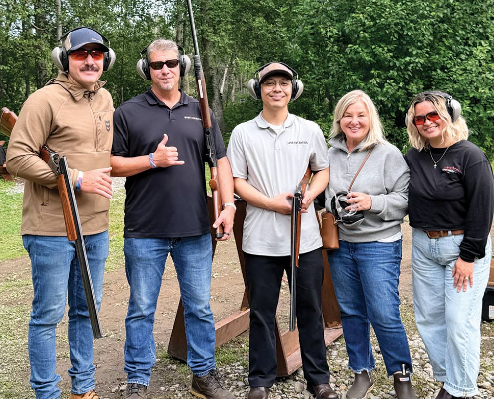 Group of five men and women wearing ear and eye protection posing with shotguns at a shooting station.
