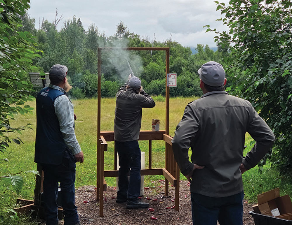 Shooter firing a shotgun from inside a wooden shooting station as two observers watch from behind.