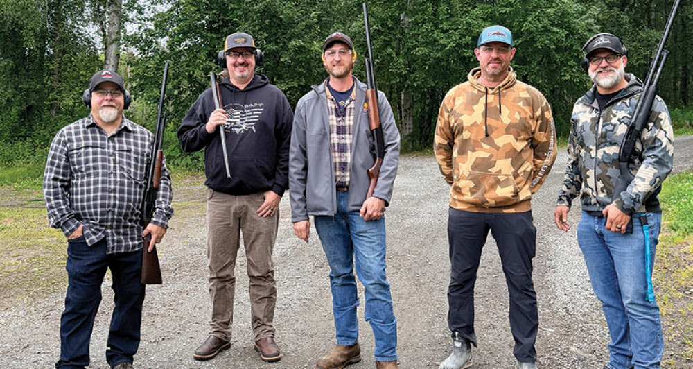 Five men in outdoor gear holding shotguns standing side by side along a gravel road.