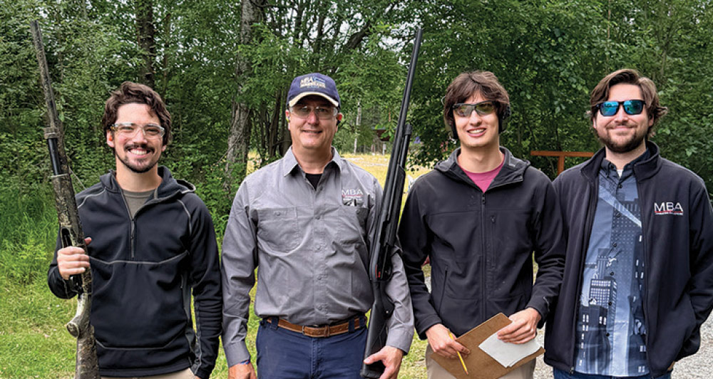 Four men wearing safety glasses and casual jackets standing together outdoors at a shooting course.