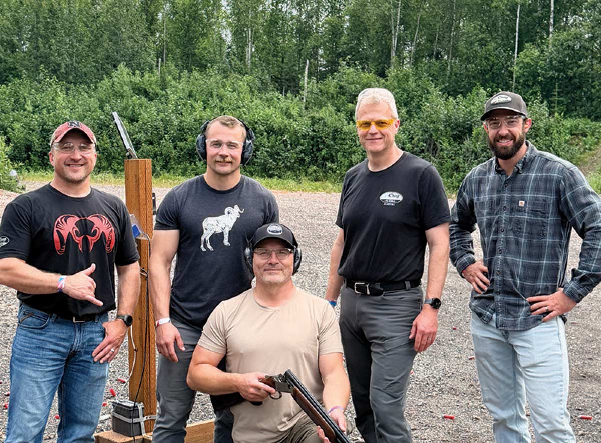 Five team members posing outdoors at a shooting station, with one kneeling while holding an open-action shotgun.