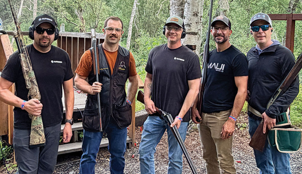 Five men in casual athletic gear holding shotguns posing together outdoors near a wooden deck.