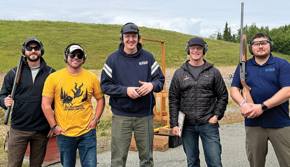 Group of five men wearing safety glasses and ear protection posing outdoors at a shooting range.