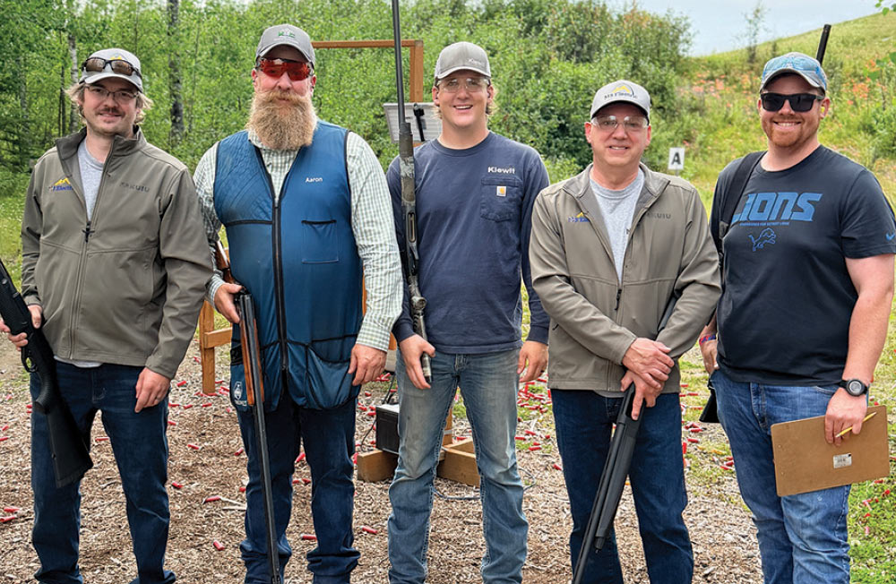 Five men in shooting gear holding shotguns standing side-by-side outdoors at a station.