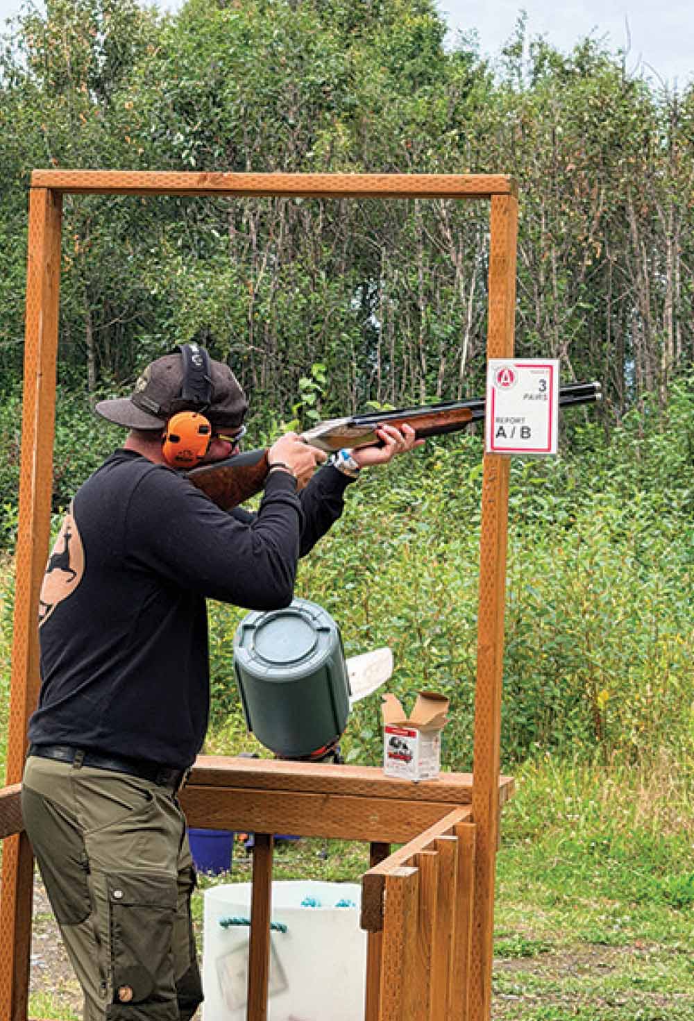 Side view of a shooter in ear protection aiming a shotgun from within a wooden shooting station.