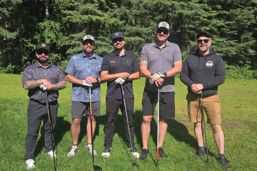 Five golfers in shorts, athletic shirts, and caps posing with their drivers in front of tall pine trees.