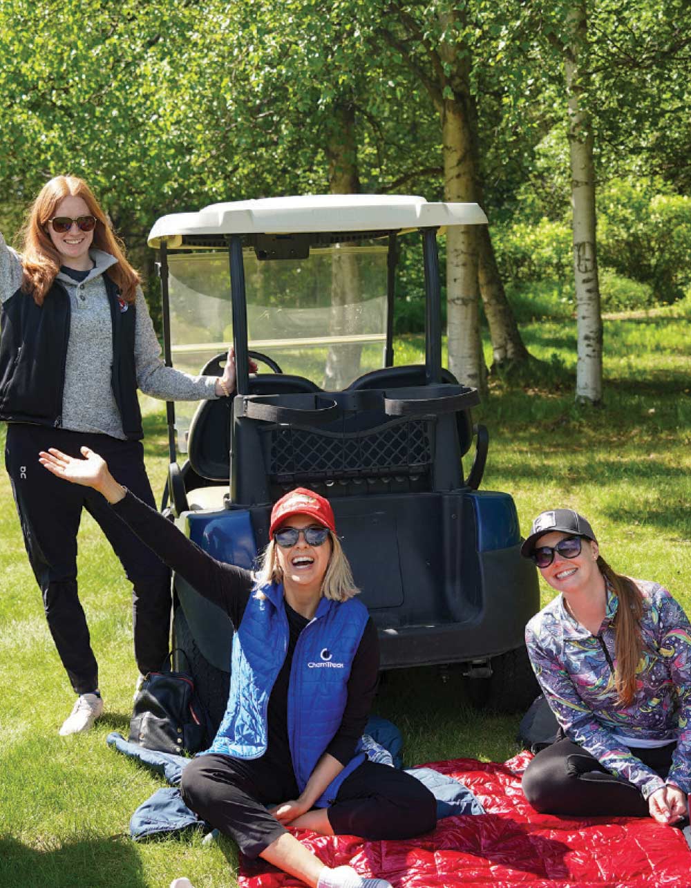 Three women smiling and posing near a golf cart, with two sitting on a red blanket on the grass.