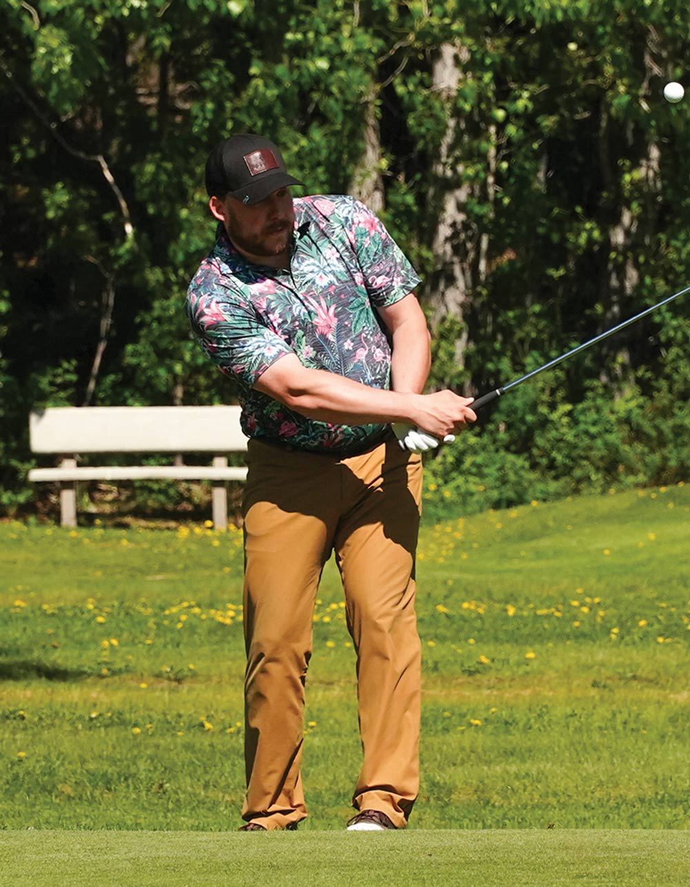 Golfer wearing a floral shirt swinging a club to hit a golf ball on a sunny day.