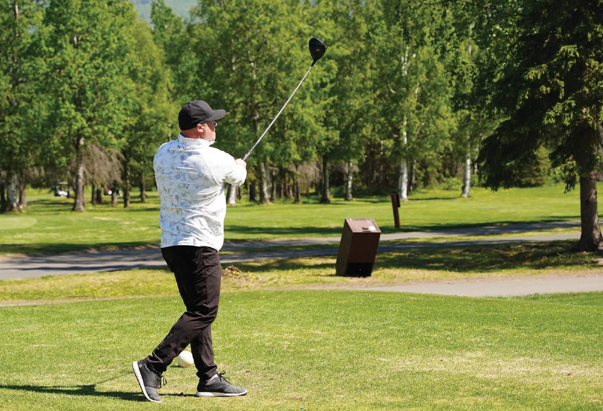 Side view of a golfer in dark pants following through on a golf swing at the tee box.