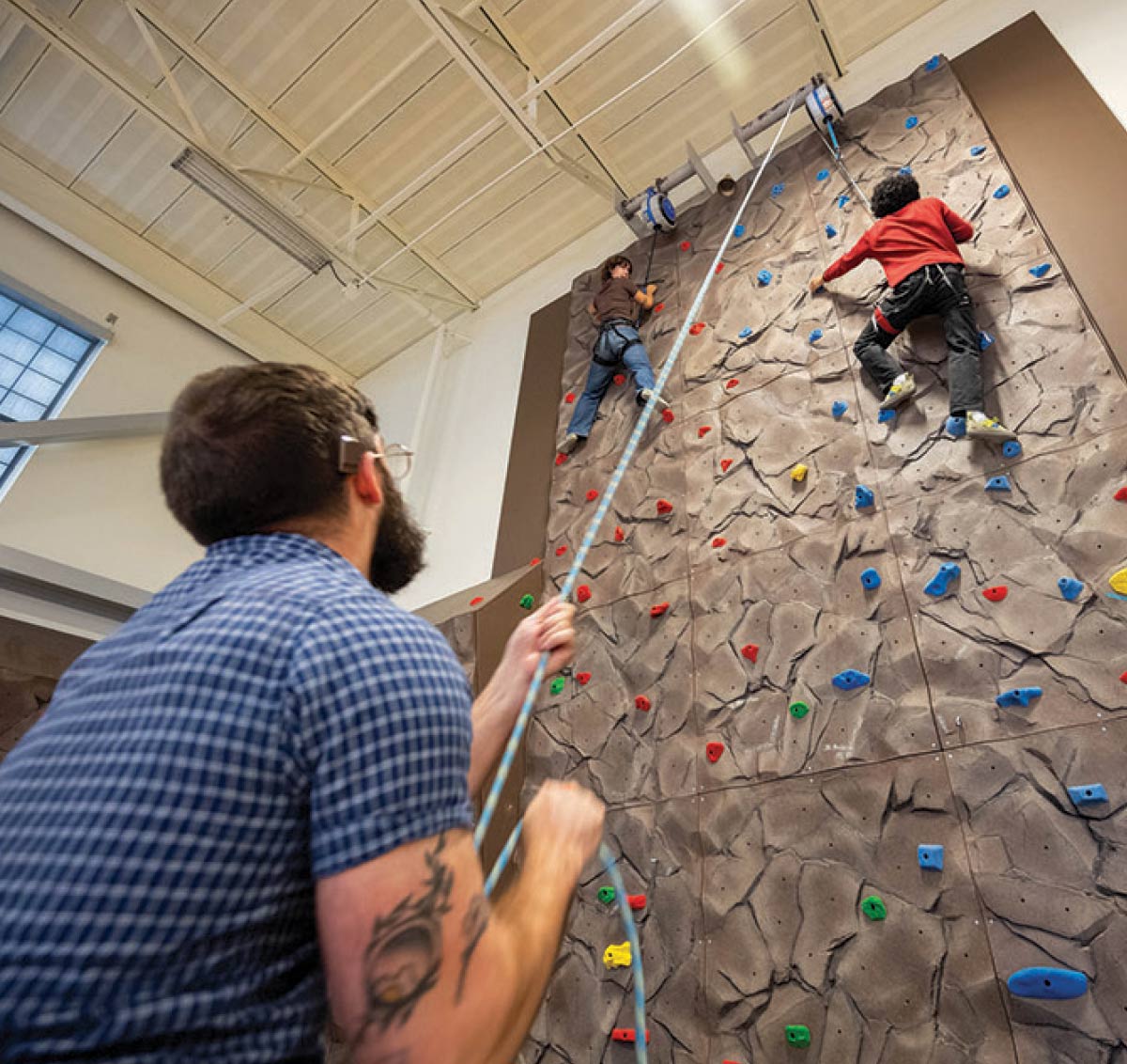 Instructor belaying a safety rope as two students scale an indoor climbing wall.