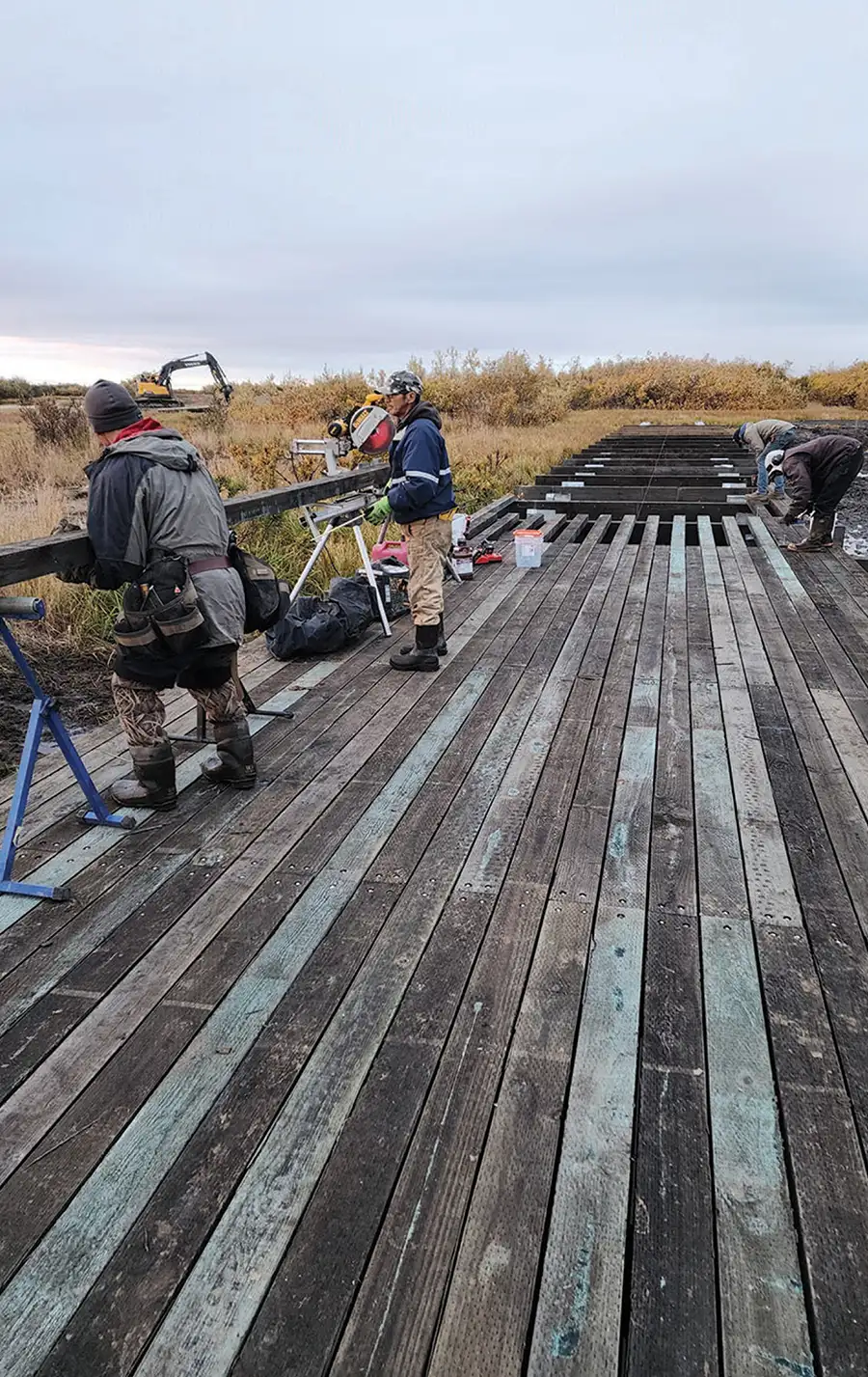 Workers building a wooden boardwalk in a rural, brushy landscape under an overcast sky. One man is using a circular saw.