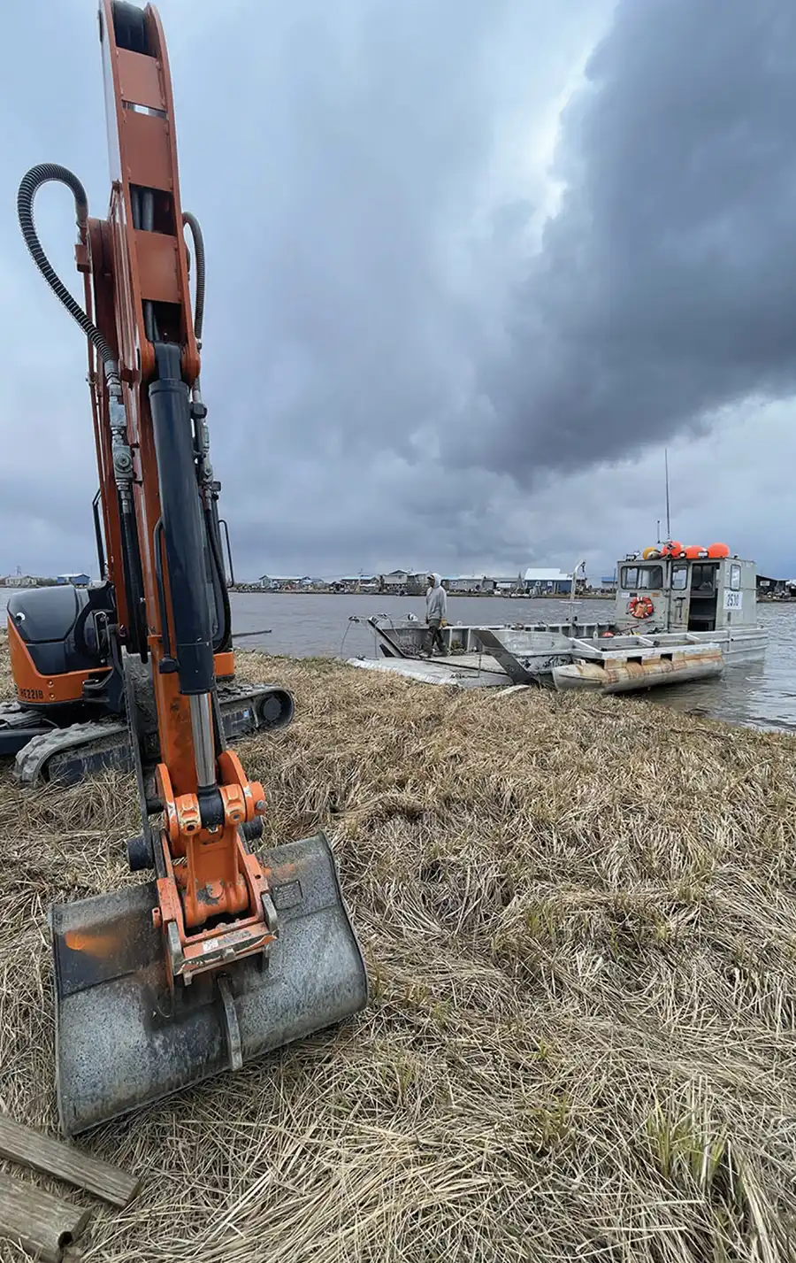 An orange excavator bucket rests on a grassy shoreline near a small landing craft boat docked at the water's edge under dark, overcast skies.