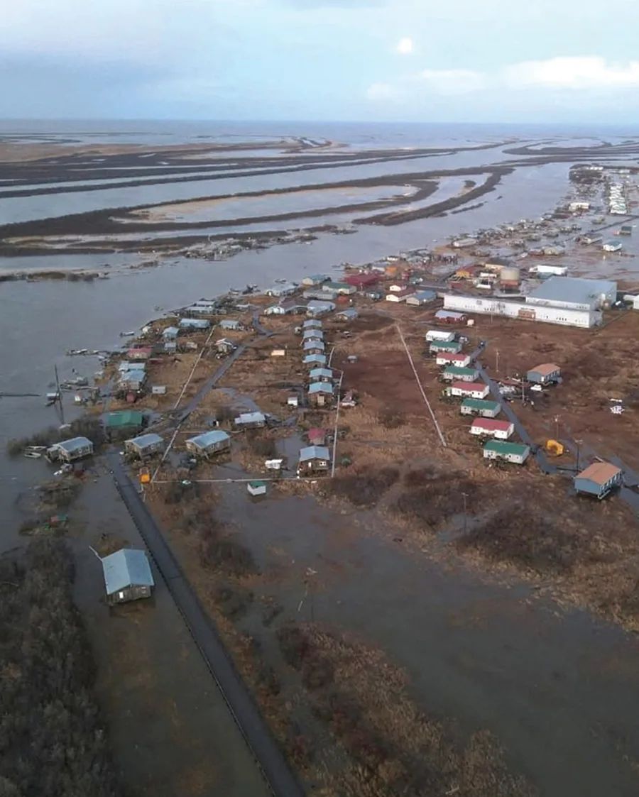 Aerial view of a rural coastal village with small homes and boardwalks, surrounded by flooded marshland and winding river delta channels.