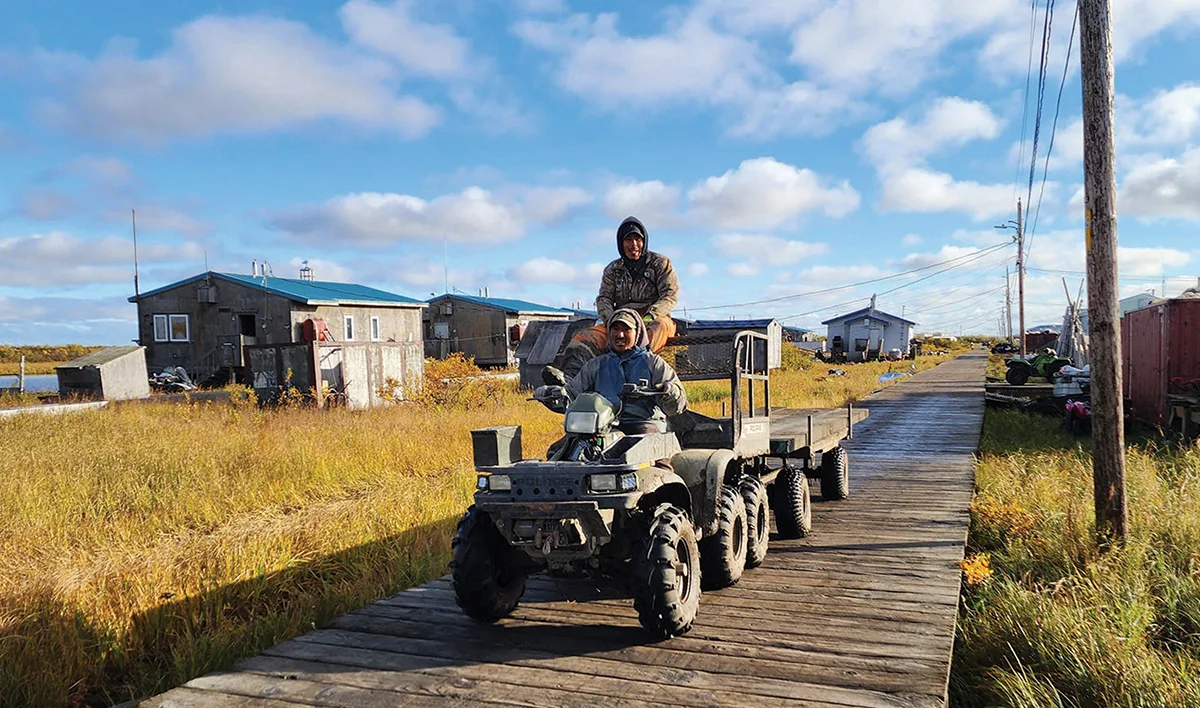 Two men ride a six-wheeled ATV towing a flatbed trailer along a wooden boardwalk through a rural village under a partly cloudy sky.