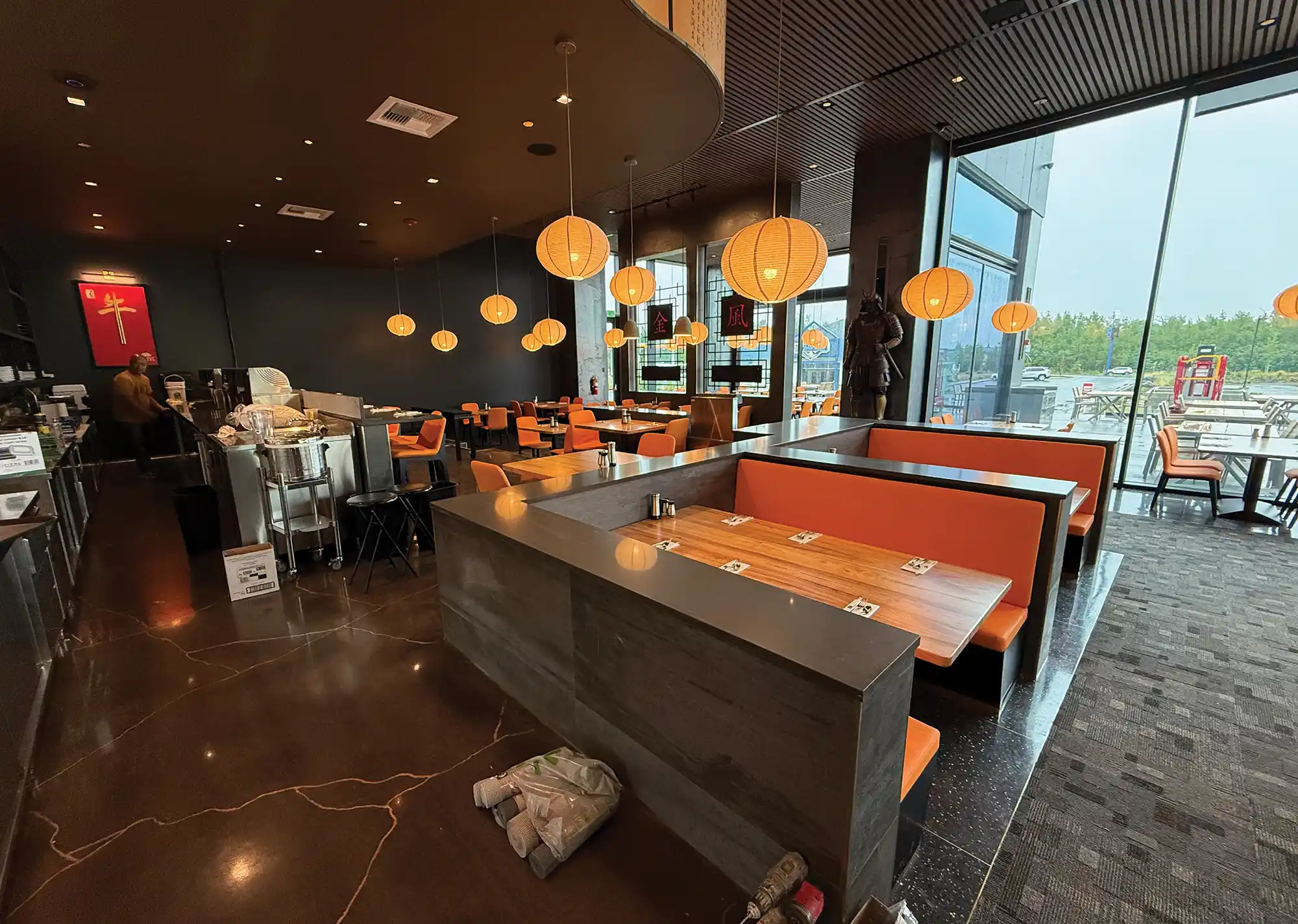 Modern restaurant interior featuring orange booth seating, wooden tables, hanging paper globe lights, dark polished floors, and floor-to-ceiling windows.