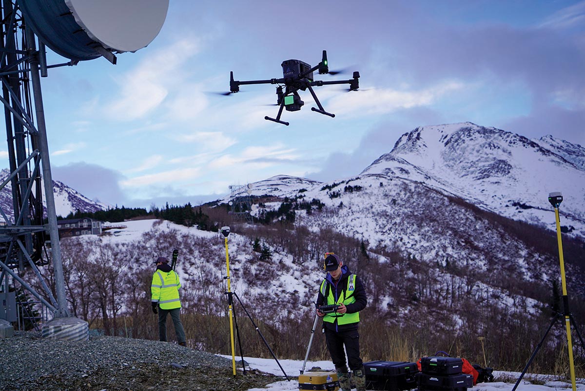 Technician operating a drone near a communications tower with snow-capped mountains in the background.