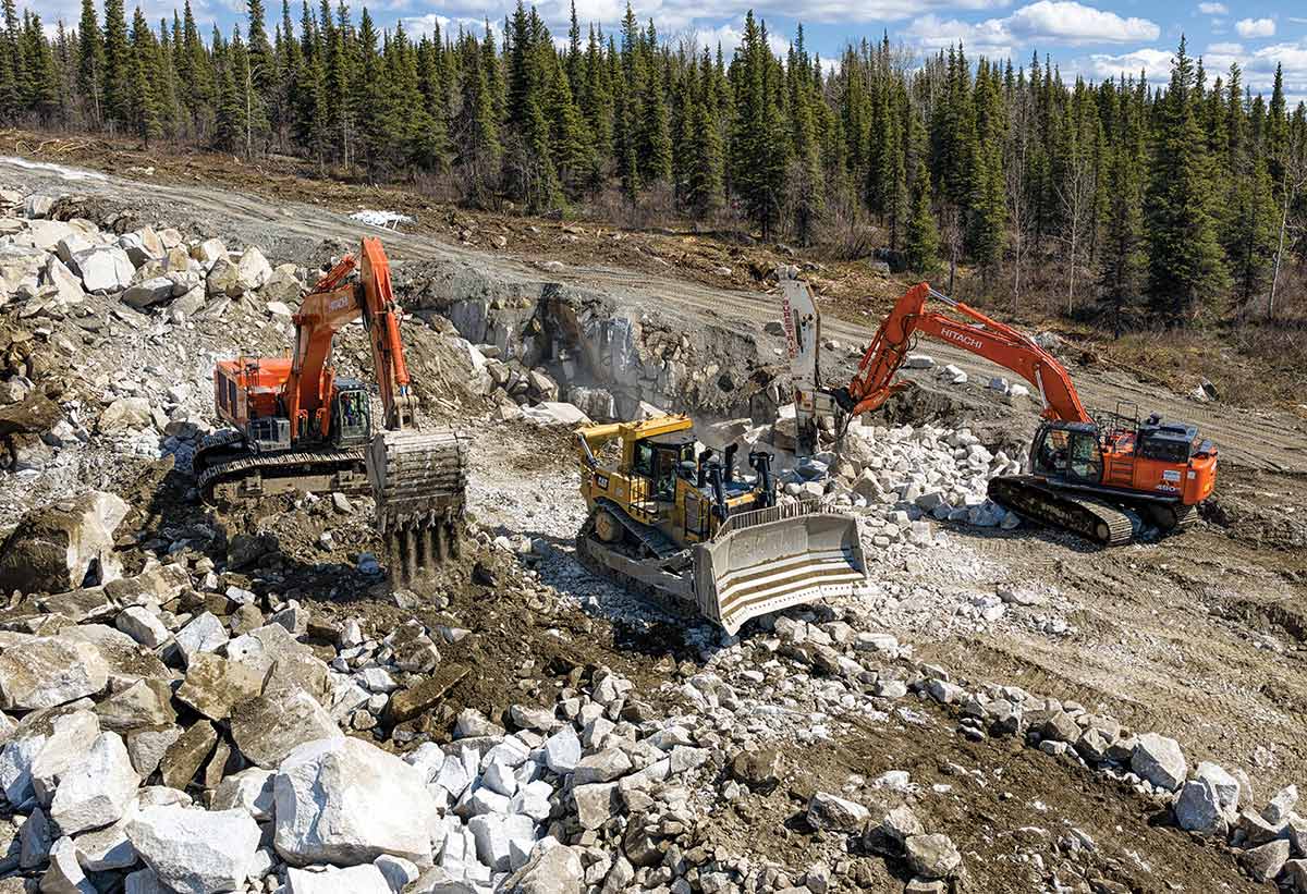 Two orange excavators and a yellow bulldozer breaking up large rocks in a forested quarry.