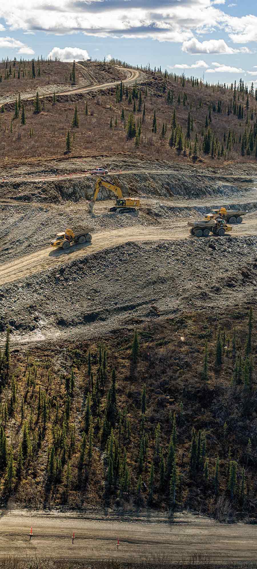 Heavy dump trucks and an excavator moving earth along a winding dirt road on a hillside.