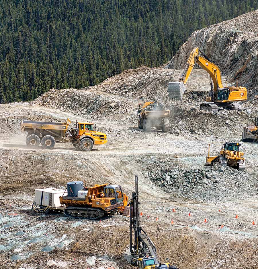 Heavy construction equipment operating at a large active excavation site along a tree line.
